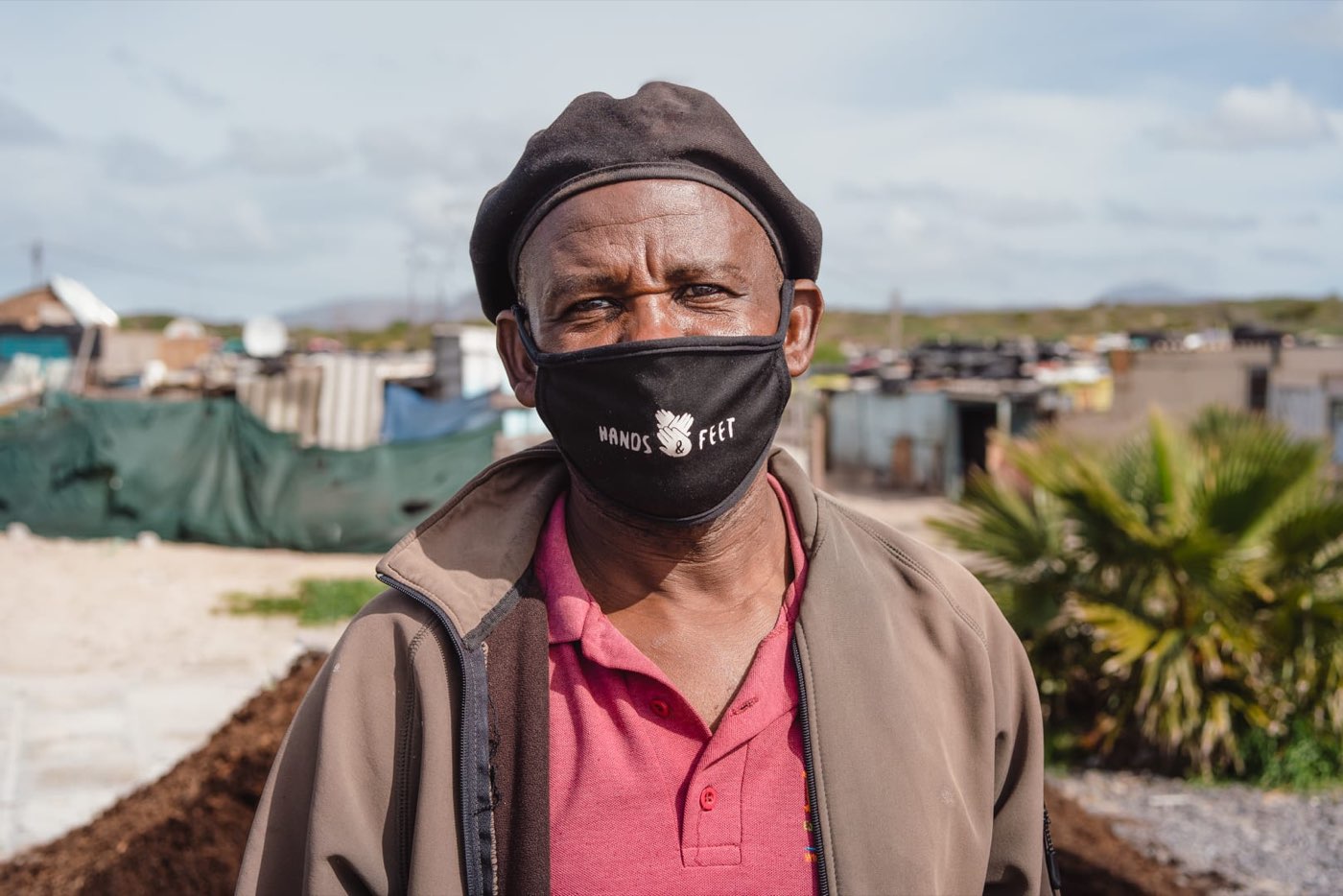 A man in a beret and face mask standing outdoors, a township behind him