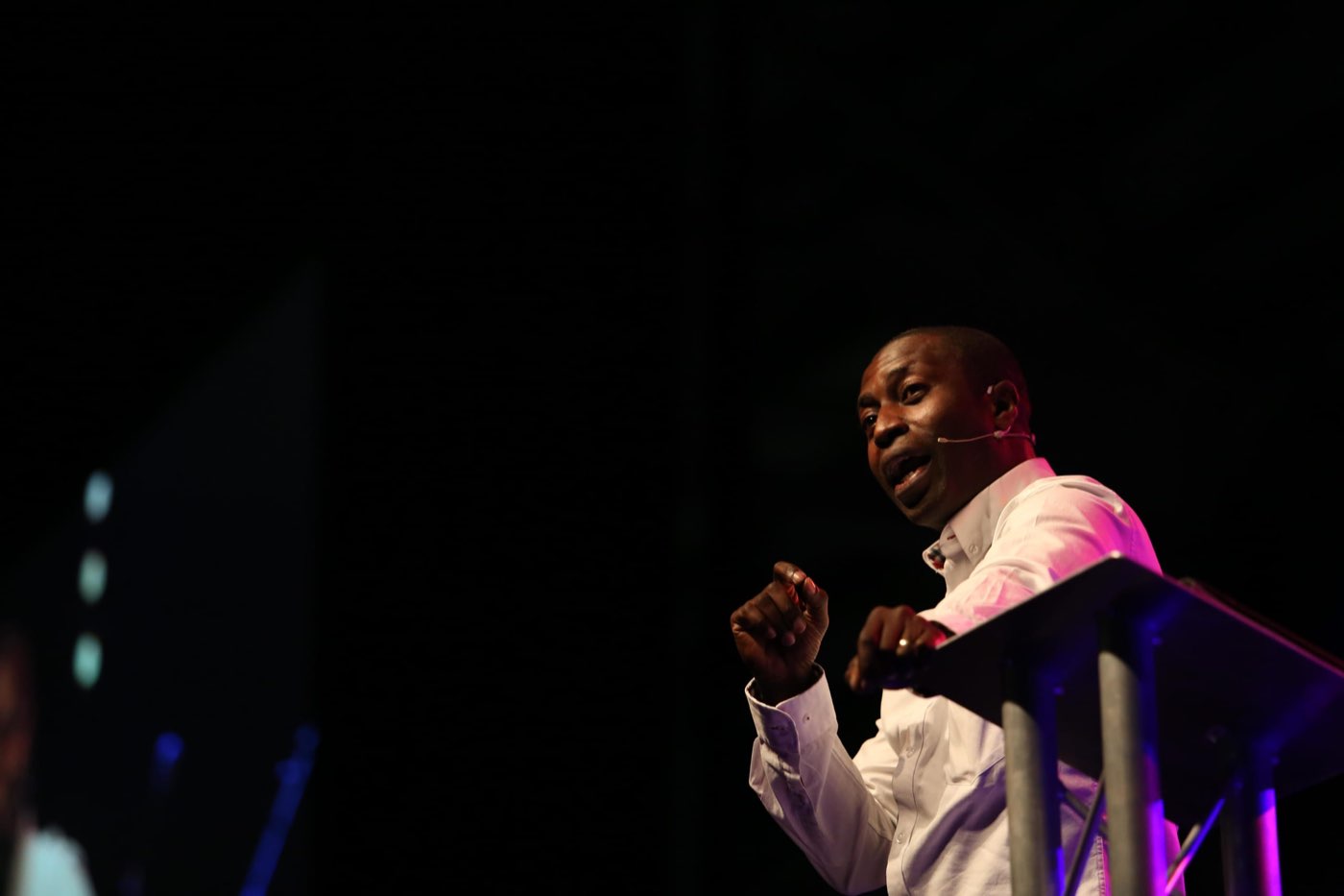 A speaker in a white shirt gesturing at a lectern on a dark stage, lit in pink
