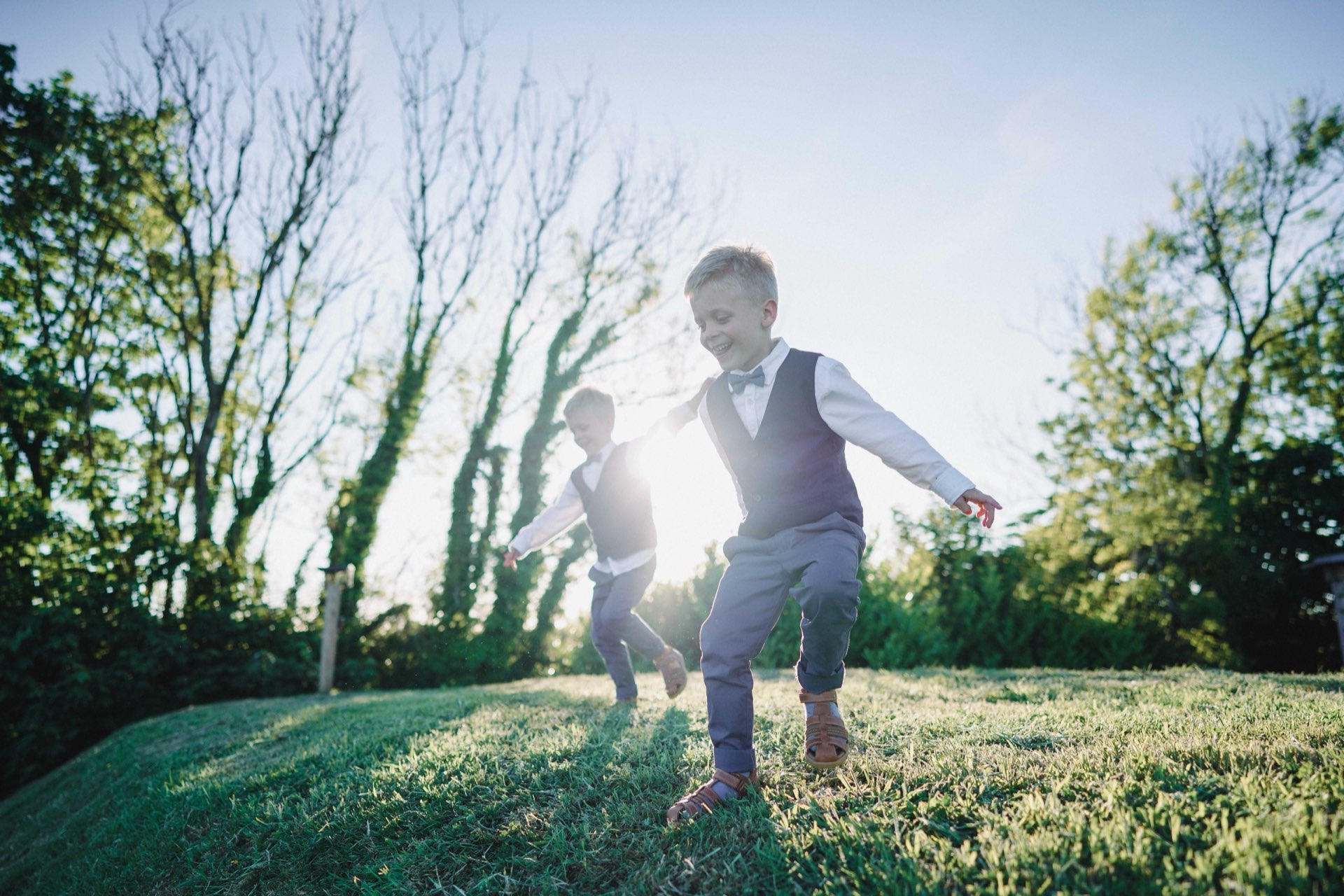 Two young page boys in smart outfits running through a sunlit garden at a wedding