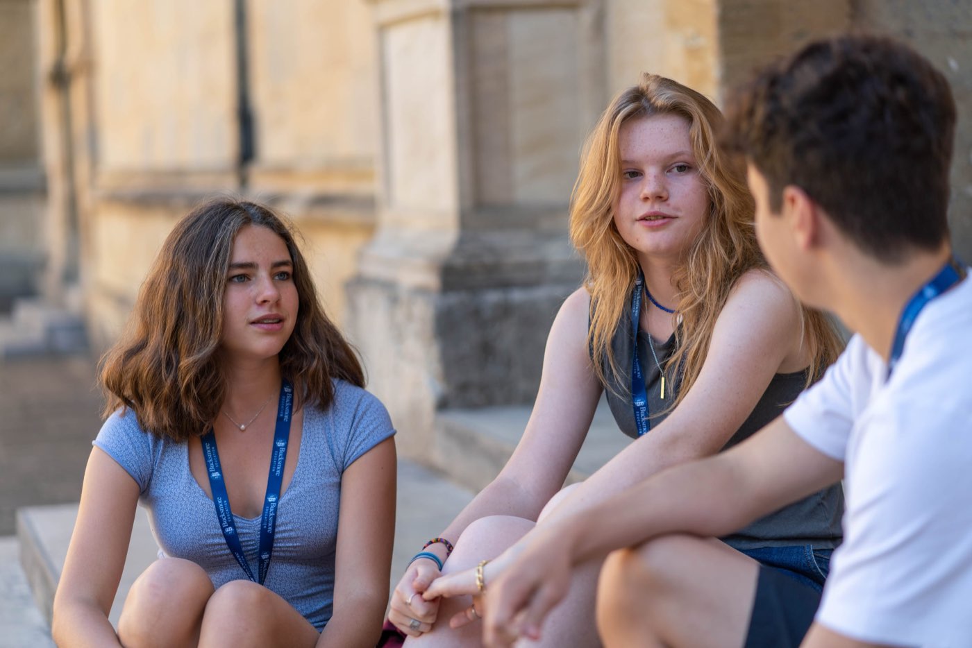 Three teenagers in lanyards sitting on stone steps, chatting