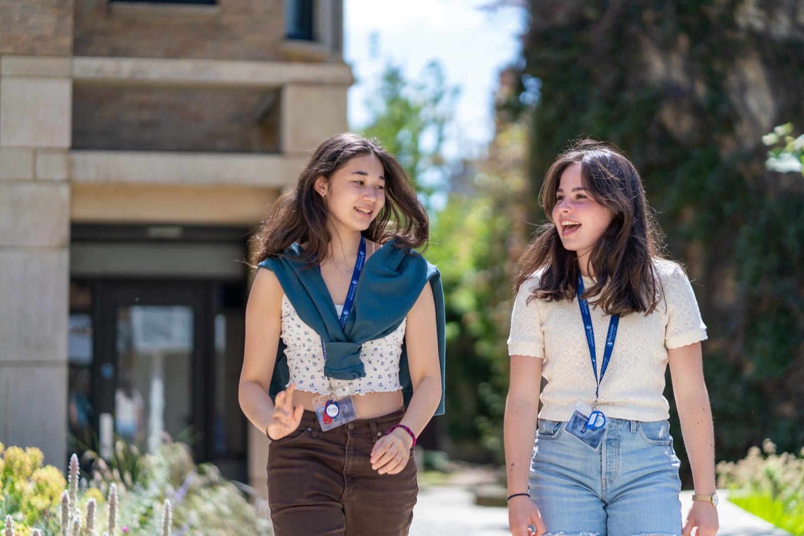 Two teenage girls in lanyards laughing as they walk past a stone building
