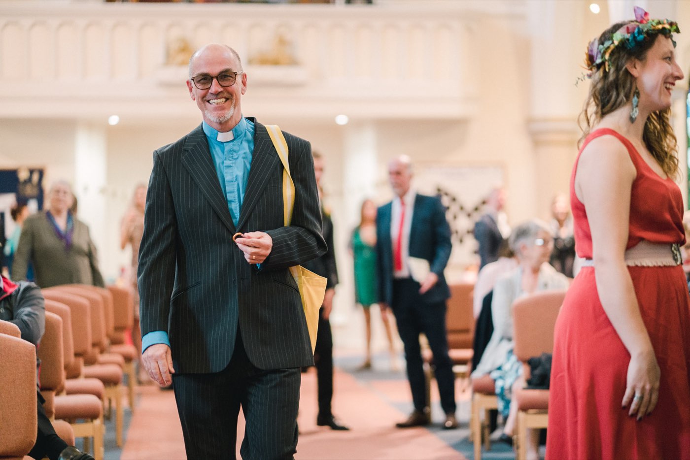 An officiant in a clerical collar smiling as he walks down the church aisle